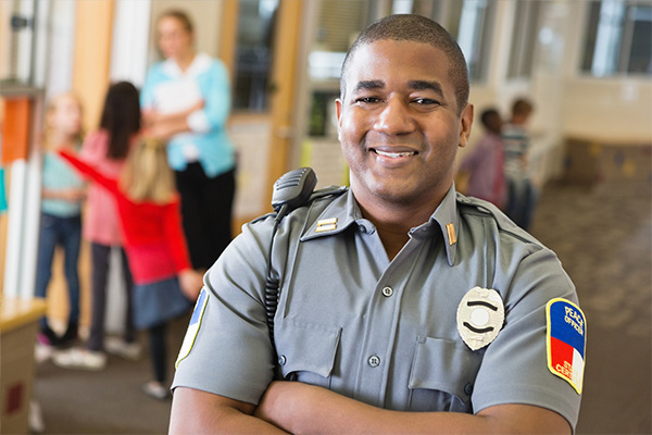 A school resource officer smiles and crosses their arms while posing for a photo in a school hallway with a teacher and children in the background
