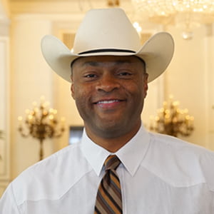 African American in white cowboy hat, white shirt, and striped tie smiles into camera