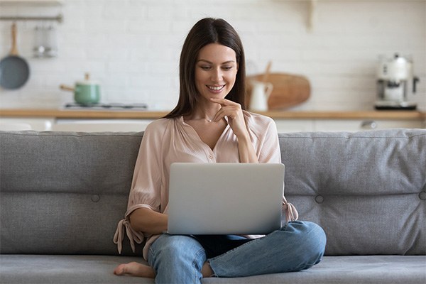 An online student smiles while sitting on a sofa looking and looking at a laptop.
