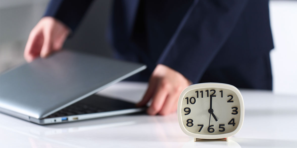 close up photo of a small clock on a desk showing 5:00 in front of hands closing a laptop